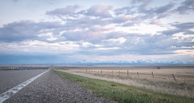 Road leading to horizon with mountains in backdrop, during sunset in Southern Alberta, Canada