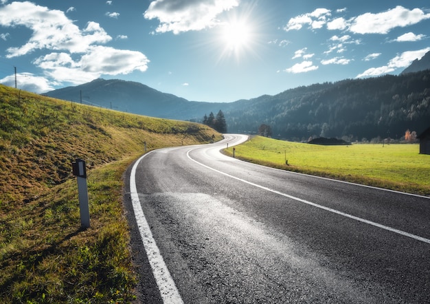 Photo road in mountain valley at sunny morning in dolomites, italy. view with asphalt roadway, meadows with green grass, mountains, blue sky with clouds and sun. highway in fields. trip in europe. travel