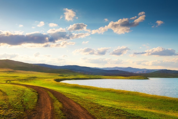 山の中の湖の近くの道路。日没時の春の自然風景。丘の上の新鮮な緑の草。南ウラル、バシコルトスタン共和国、ロシア。