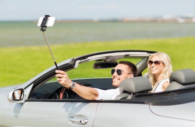 도로 여행, 레저, 커플, 기술 및 사람 개념 - 행복한 남자와 여자가 카브리올레 차를 타고 셀카봉에 스마트폰으로 사진을 찍는다