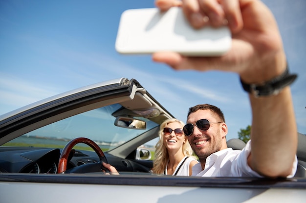 road trip, leisure, couple, technology and people concept - happy man and woman driving in cabriolet car and taking selfie with smartphone