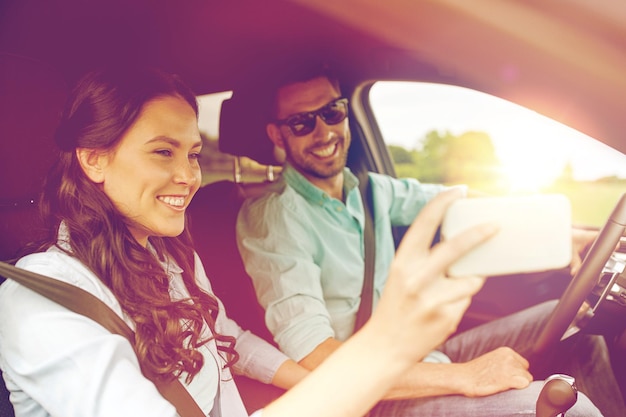 road trip, leisure, couple, technology and people concept - happy man and woman driving in car and taking selfie with smartphone