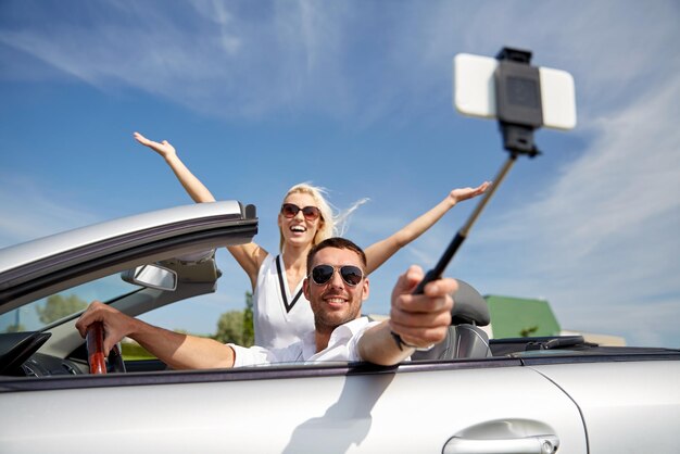 road trip, travel, couple, technology and people concept - happy man and woman driving in cabriolet car and taking picture with smartphone on selfie stick