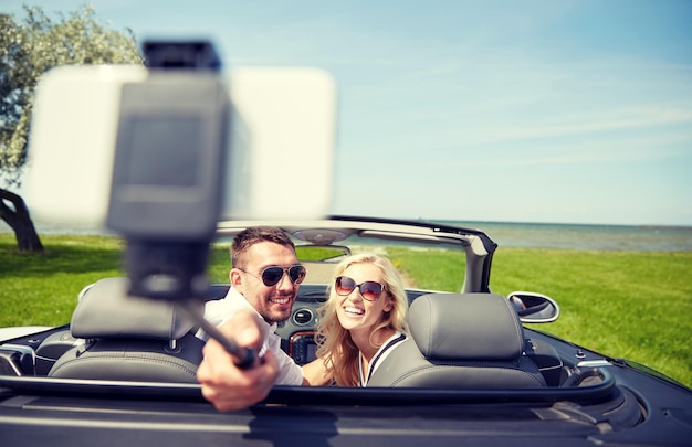 путешествие, путешествие, пара, технология и концепция людей - счастливый мужчина и женщина за рулем кабриолета и фотографируются со смартфоном на селфи-палке
