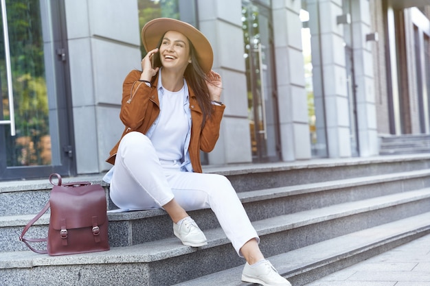 と電話で話している帽子とバックパックを身に着けている若い幸せな女性旅行者や観光客を歩き回る