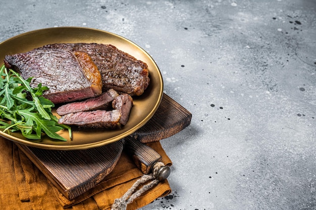 Roast picanha or top sirloin beef meat steak on a plate with arugula salad Gray background Top view Copy space