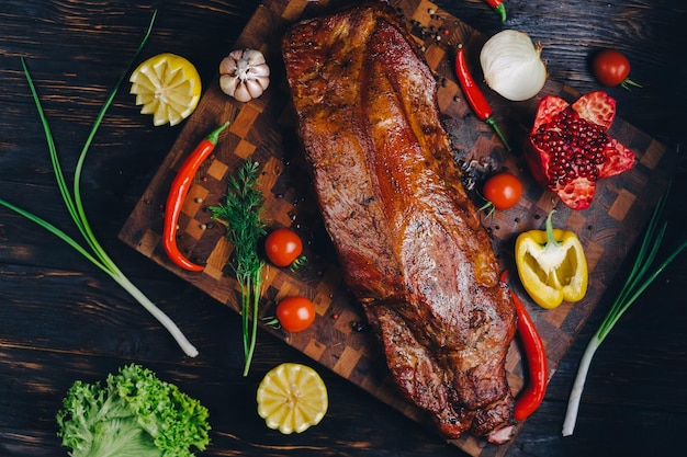 Photo roast pork in a smokehouse standing on a cutting board garnished with rosemary pepper chili pepper tomato lemon pomegranate aromatic baked meat