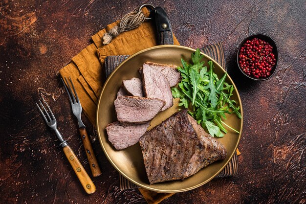 Roast Tri Tip or sirloin bottom beef steak on a plate with arugula. Dark background. Top view.