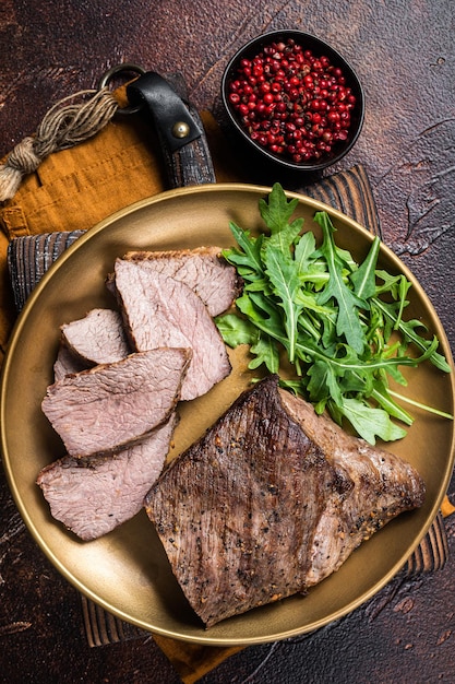 Roast Tri Tip or sirloin bottom beef steak on a plate with arugula Dark background Top view