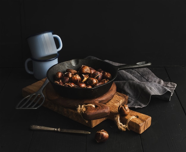 Roasted chestnuts in skillet cooking pan over rusti wooden boards, blue enamel mugs, towel on dark 