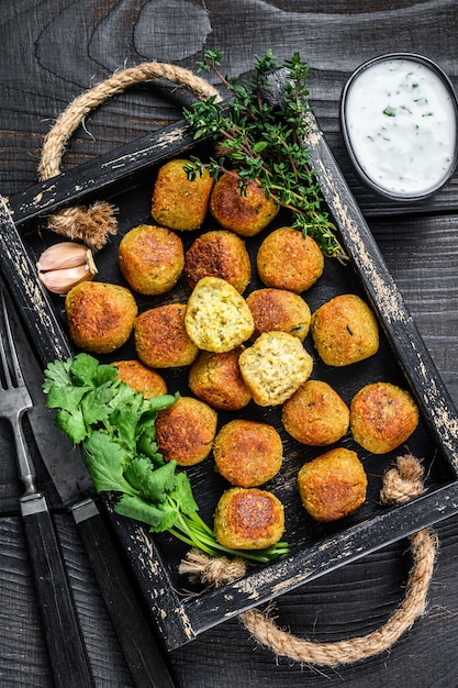 Roasted chickpeas falafel balls with garlic yogurt sauce in a wooden tray. Black wooden background. Top view.