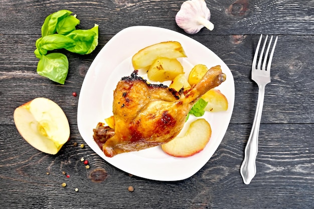 Жареная утиная ножка с яблоком, картофелем в белой тарелке, базиликом, чесноком и вилкой на фоне деревянных досок сверху