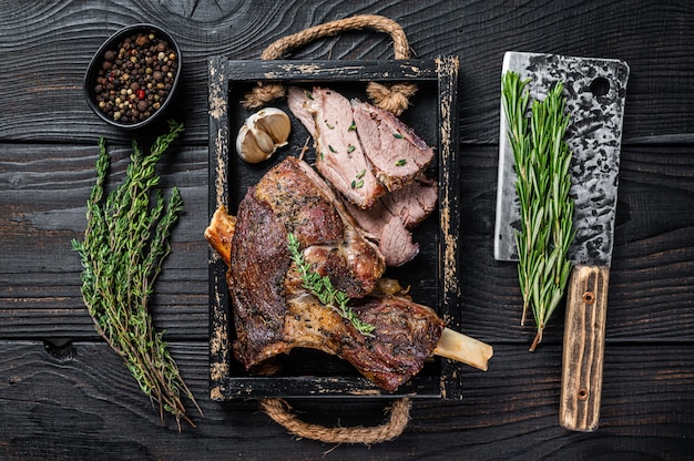 Roasted mutton lamb leg sliced in a wooden tray with meat\
cleaver. black wooden table. top view.