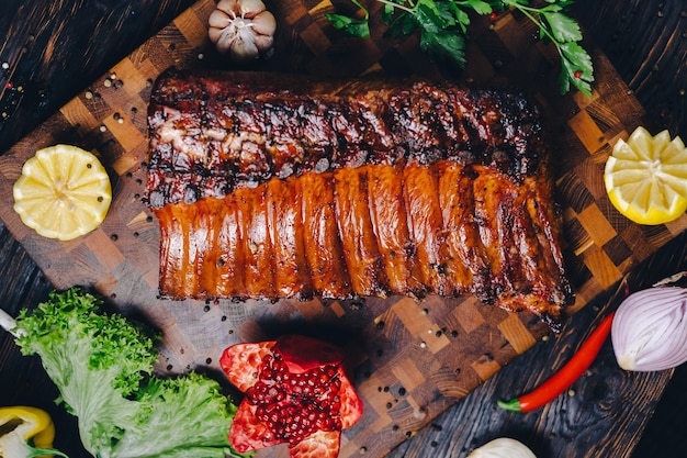 Photo roasted pork ribs in a smokehouse standing on a cutting board garnished with rosemary pepper chili pepper tomato lemon pomegranate aromatic baked meat