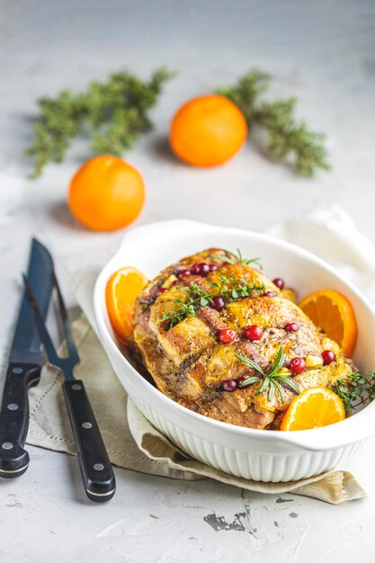 Roasted pork in white dish christmas baked ham with cranberries tangerines thyme rosemary garlic on light table surface