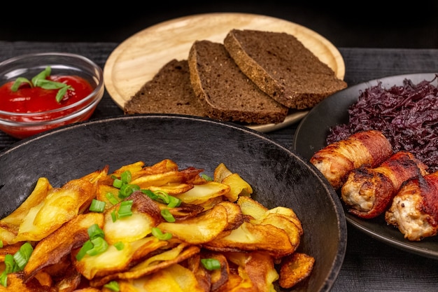 Roasted potato in the pot, pieces of rye black bread on a wooden serving plate, homemade sausages in bacon and stewed cabbage on a black rustic background.