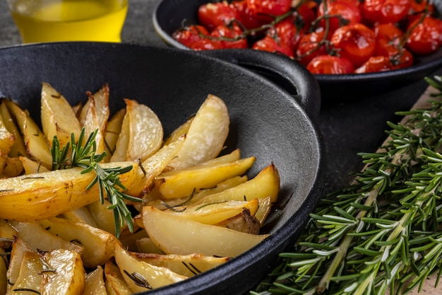 Roasted potatoes with rosemary in iron casserole and plate of confit tomatoes on dark background.