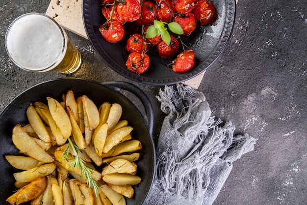 Roasted potatoes with rosemary in iron casserole and plate of confit tomatoes on dark surface.