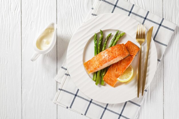 Roasted salmon fillets with grilled asparagus and lemon wedges on a white plate on a white wooden table with hollandaise sauce horizontal view from above flat lay