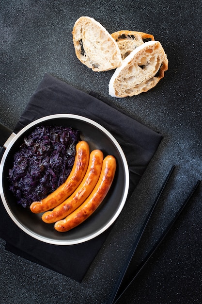 Roasted sausages bratwurst served in pan with bread and red cabbage on a black background. Traditional German dish, German cuisine. Top view, copy space