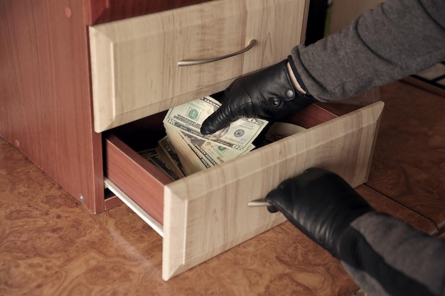 Robber in black outfit and gloves see in opened shelf in\
kitchen the thief takes out the us dollar bills from a shelf