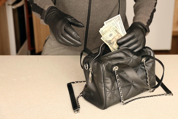 Robber in black outfit and gloves see in opened stolen women\
bag the thief takes out us dollar bills from a womans handbag in\
kitchen