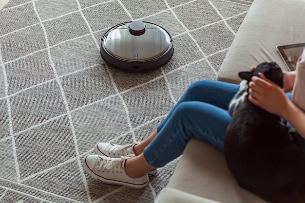 Robot vacuum cleaner cleaning carpet, woman and cat rest sitting on the sofa at home