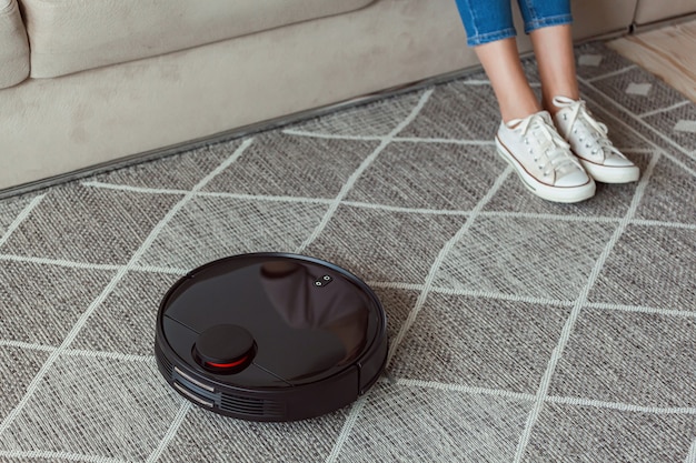 Робот-пылесос чистит ковер, ноги женщины отдыхают, сидя на диване у себя дома