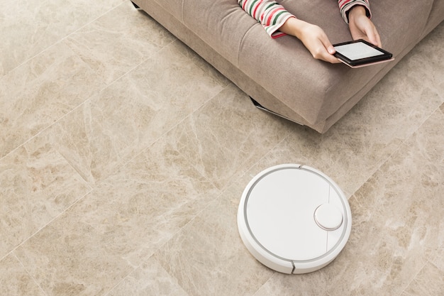 Robot vacuum cleaner cleaning the room while woman relaxing on sofa