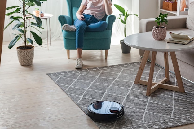 Robotic vacuum cleaner cleaning carpet woman controls remote control and joy rest while sitting on armchair at home