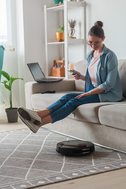 Робот-пылесос чистит ковер, женщина дистанционно управляет мобильным телефоном и наслаждается отдыхом, сидя на диване у себя дома