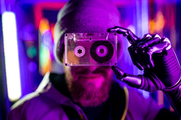Фото Человек-робототехник с металлической рукой ностальгирует о прошлом в неоновой студии, чувствуя одиночество, держит аудиокассету