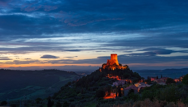 Рокка д'Орча - средневековая деревня и крепость в долине Орча, Тоскана, Италия. Уникальный вид в сумерках на каменную башню на скале, освещенную на фоне драматического неба.