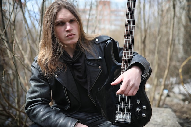 Photo rock guitarist on the steps. a musician with a bass guitar in a leather suit. metalist with a guitar on the background of industrial step.