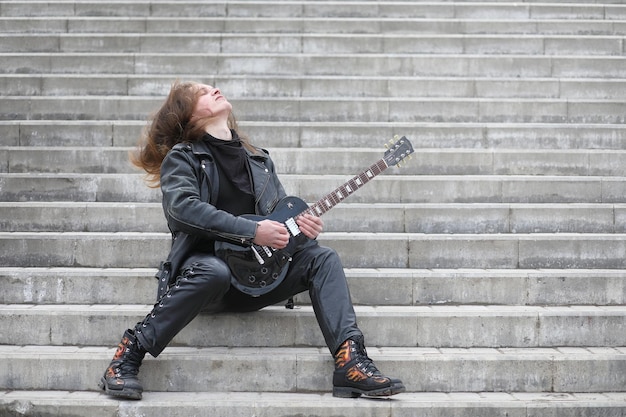 Photo rock guitarist on the steps a musician with a bass guitar in a leather suit metalist with a guitar on the background of industrial steps