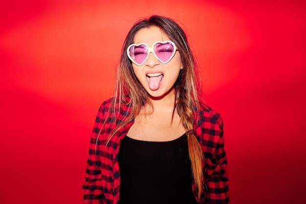 Foto rock'n'roll ragazza kazaka in una camicia a quadri su sfondo rosso mostra giocosamente la lingua negli occhiali a forma di cuore, ritratto in studio
