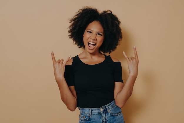 Photo rock star. young self confident afro american woman holding both hands in rock n roll sign and shouting loudly with emotional face expression, feeling carefree while posing against beige background