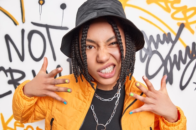 Rock style and urban lifestyle concept. Cheeky woman smirks face raises hands enjoys rap music dressed in fashionable outfit poses against graffiti wall