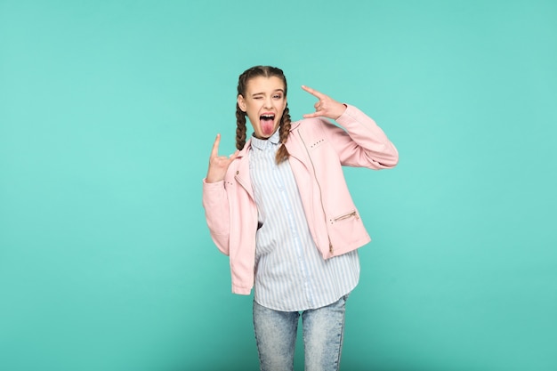 Rock tongue out portrait of beautiful cute girl standing with makeup and brown pigtail hairstyle in striped light blue shirt pink jacket. indoor, studio shot isolated on blue or green background.