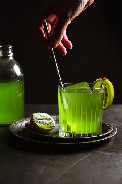 Фужер с освежающим зеленым напитком и кубиком льда внутри, украшенный киви, бутылка. Смешивание напитка барной ложкой.