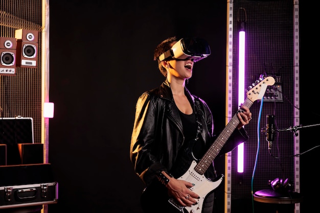 Rockstar with vr goggles enjoying rock concert simulation while playing heavy metal song at electric guitar in music studio. Woman musician performing grunge album using electricinstrument