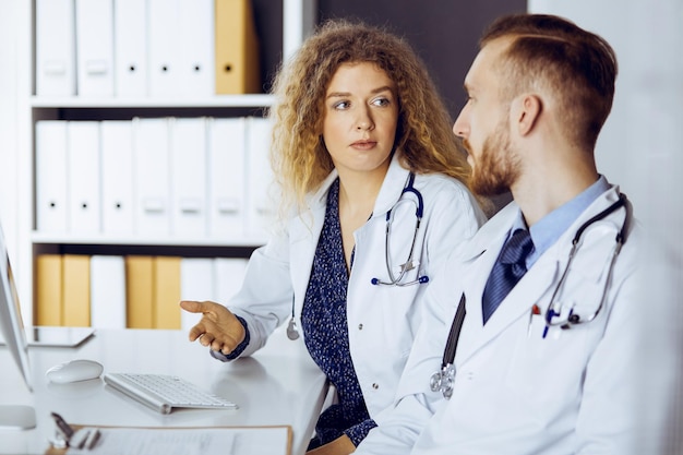 Rode bebaarde arts met vrouwelijke collega die de huidige ziektetherapie bespreekt terwijl hij op de werkplek in de kliniek zit. Teamwerk in de geneeskunde.