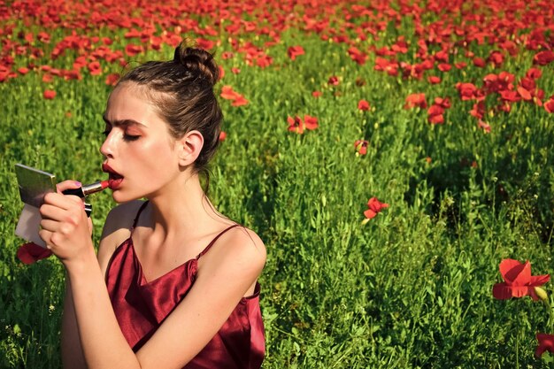 Rode lippen van mooie vrouw zetten make-up op met spiegel en lippenstift Jonge vrouw in papaverbloemveld yout