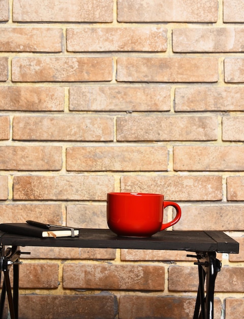 Rode thee- of koffiekop of keramische mok voor drank-exemplaar en pen zwart dienblad op baksteen getextureerde achtergrondkopieerruimte