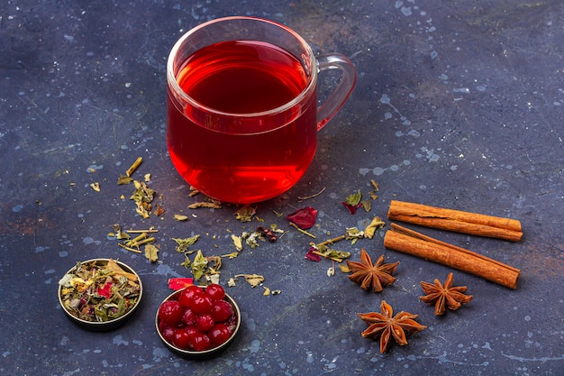 Rode thee (rooibos, hibiscus, karkade) in glazen beker en theepot onder kaneel, anijs, veenbessen op een donkere achtergrond. Kruiden, vitamine, detox thee voor verkoudheid en griep. Close-up, kopieer ruimte voor tekst