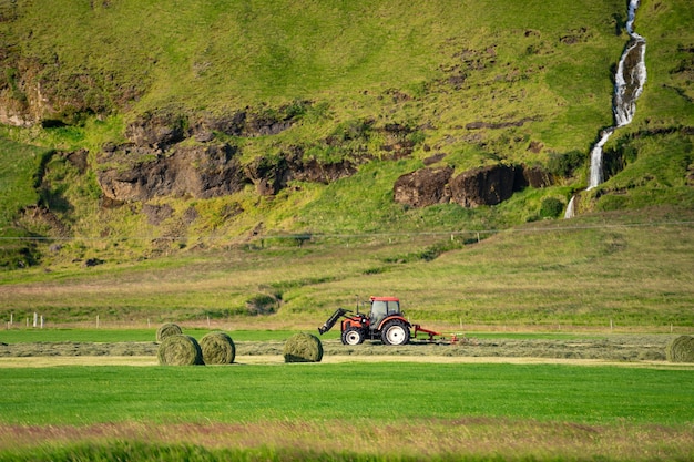 Rode trekker gras verzamelen op groen veld met prachtige rivier gaan door de berg op achtergrond.