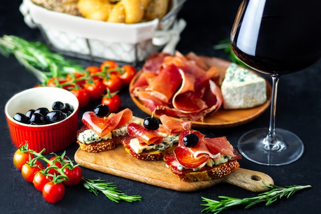 Rode wijn wordt gegoten in een bel kaas, jamon, prosciutto en olijven op een zwarte achtergrond. Wijnsnack op een houten bord. Brood met kaas en wijnsnacks. Heerlijke snacks voor het feest.