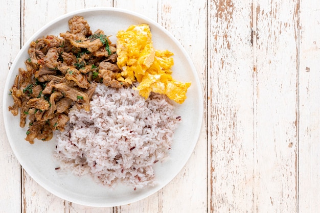 Foto roergebakken rundvlees met boombasilicumblaadjes, lange peper, indiase lange peper, javaanse lange peper, peper, knoflook, chili naast roerei en gestreamde rijst op witte houtstructuurachtergrond, bovenaanzicht