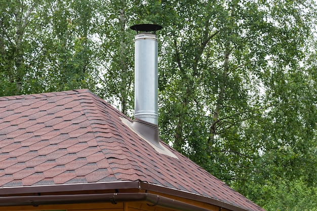 Roestvrijstalen metalen schoorsteenpijp op het dak van het huis tegen groene boom.