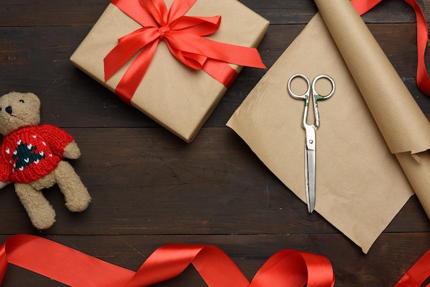 Roll of brown kraft packaging paper, box tied with a red silk ribbon, scissors and a bobbin with ribbon on a brown wooden background, top view